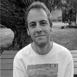 A young man with short hair and a slight smile, wearing a light-colored T-shirt, standing outdoors next to a large tree with a park in the background.