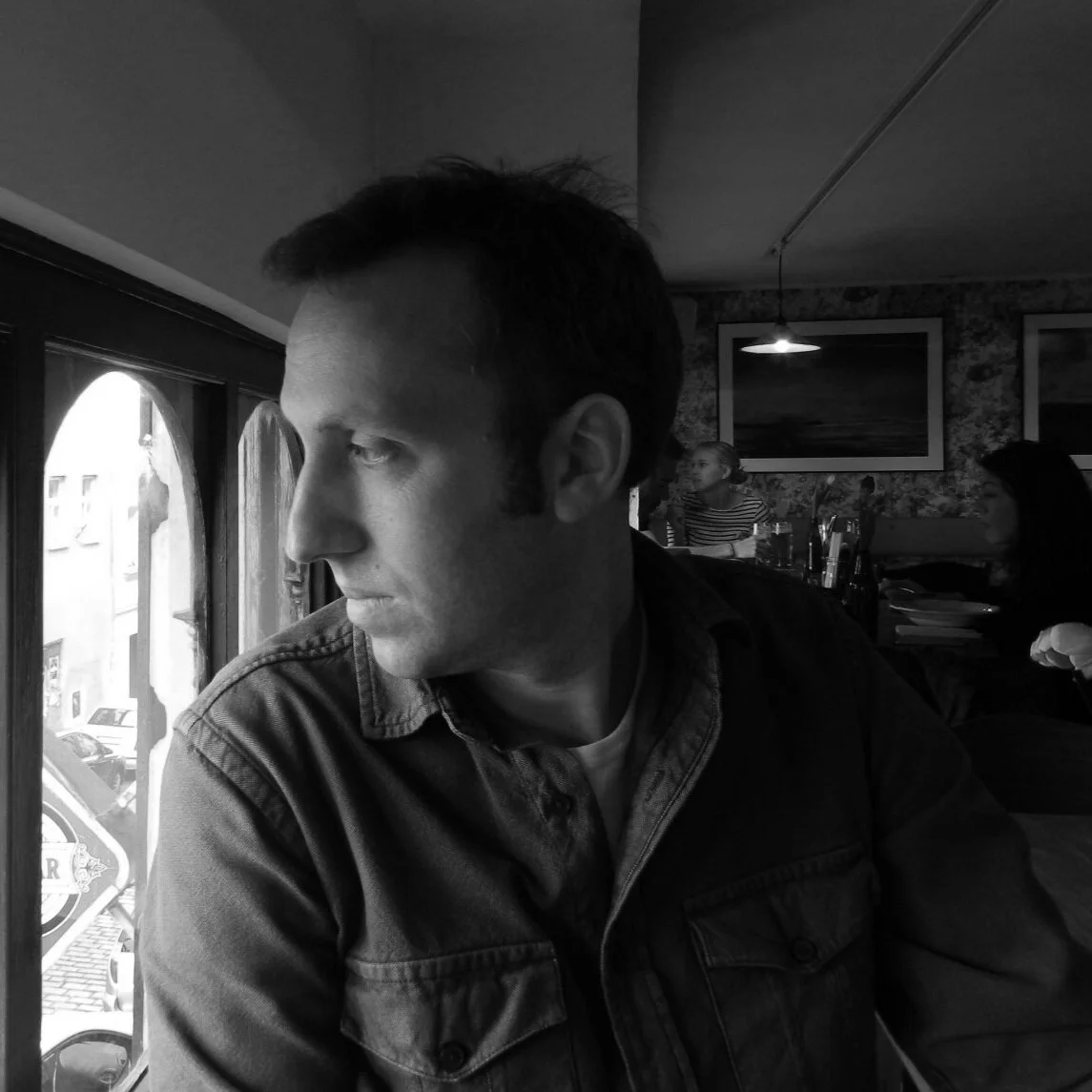 A black and white photo of a man looking out a window in a restaurant or cafe, with two women in the background, one wearing a striped shirt and the other with dark hair, sitting at a table.
