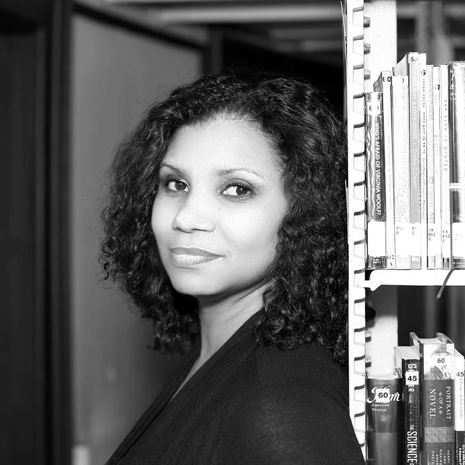 Portrait of a woman with curly hair standing next to a bookshelf in a library, looking at the camera.