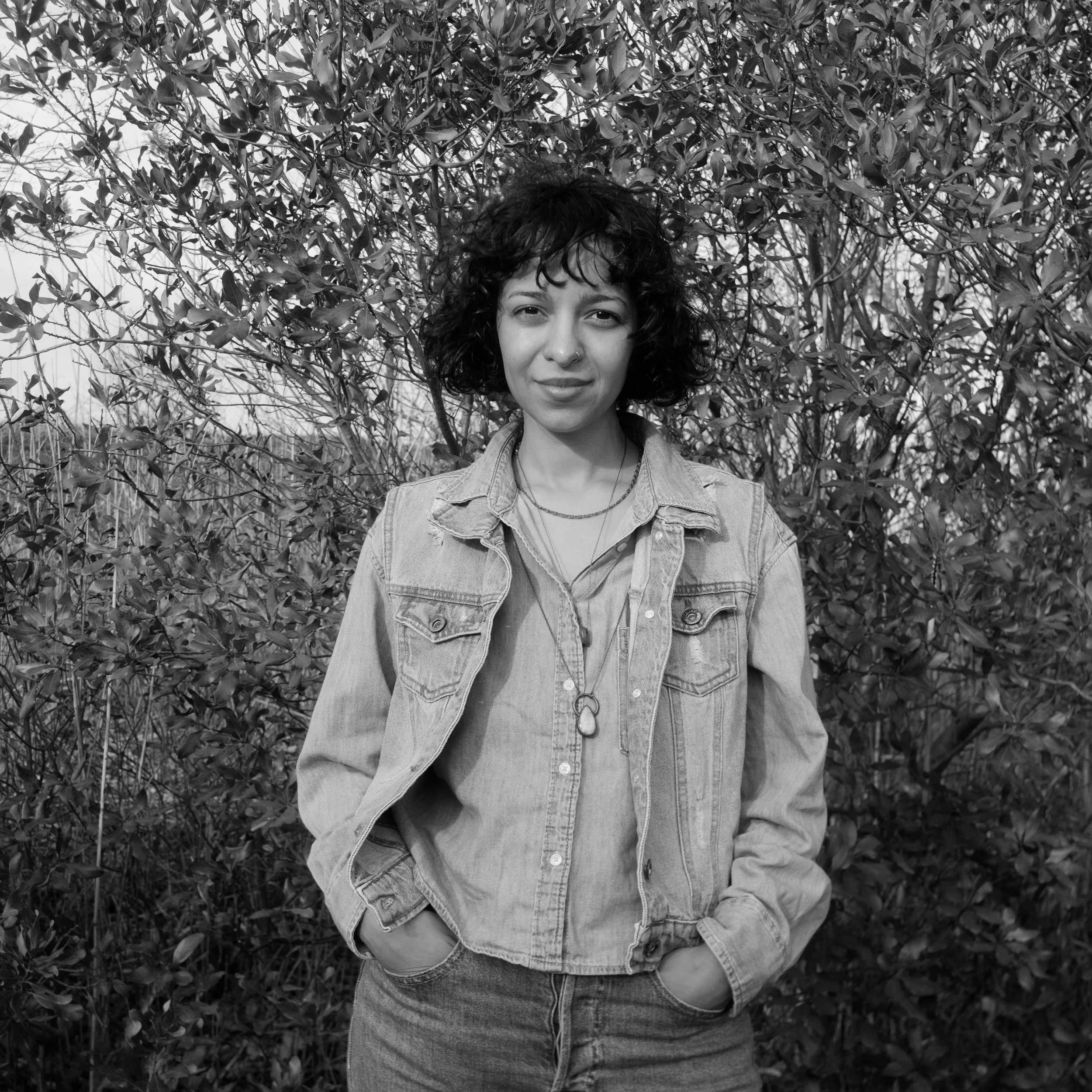 A young woman with curly hair, wearing a denim jacket and jeans, standing outdoors in front of a bush with dense leaves.