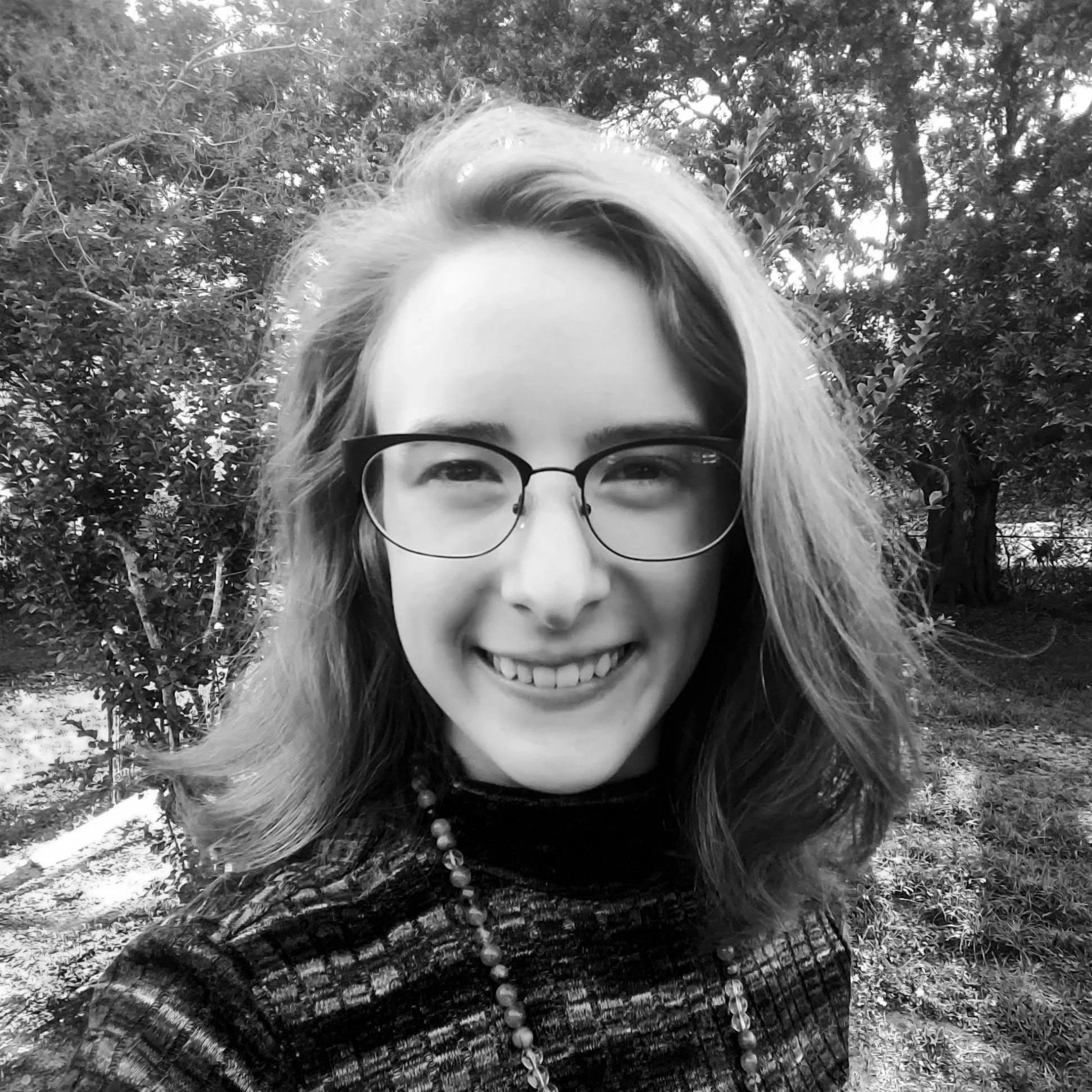 A smiling woman with glasses and wavy hair outdoors in front of trees.