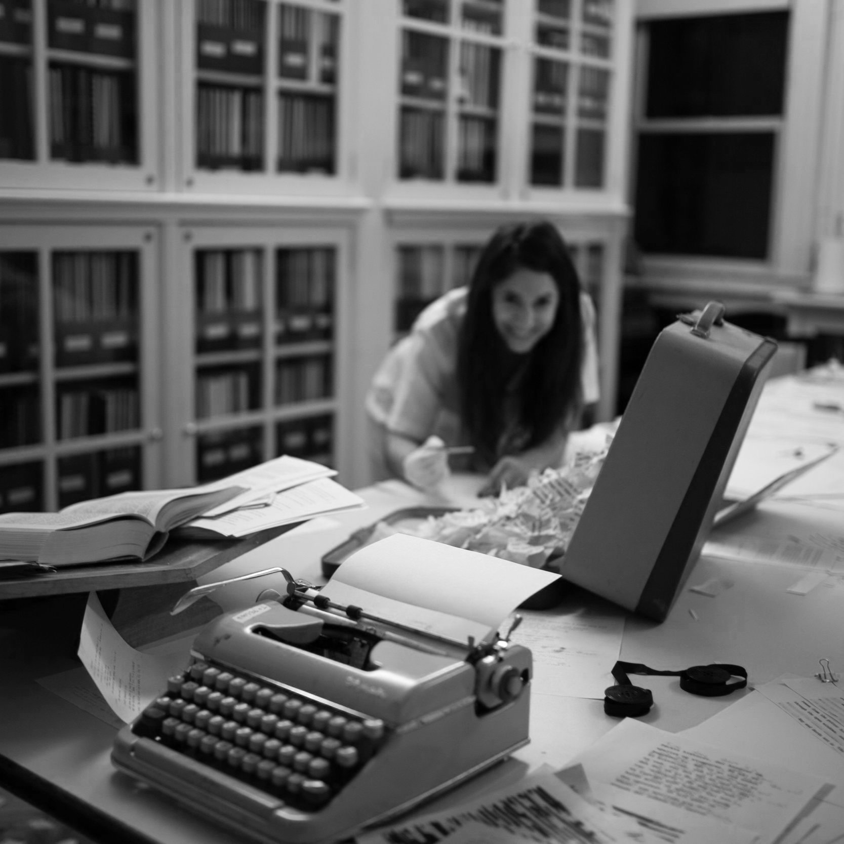 A woman smiling at a desk cluttered with papers, an open book, a typewriter, a slice of crumpled paper, and a tape recorder, in a room with shelves filled with files.