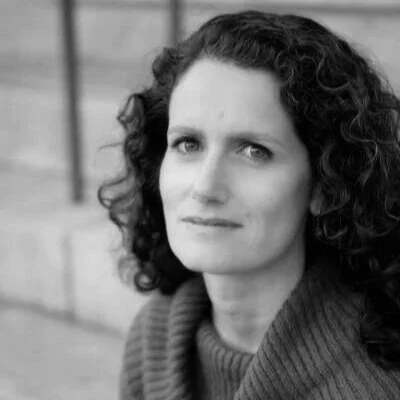 A woman with curly hair wearing a sweater, sitting outdoors with wooden stairs in the background.