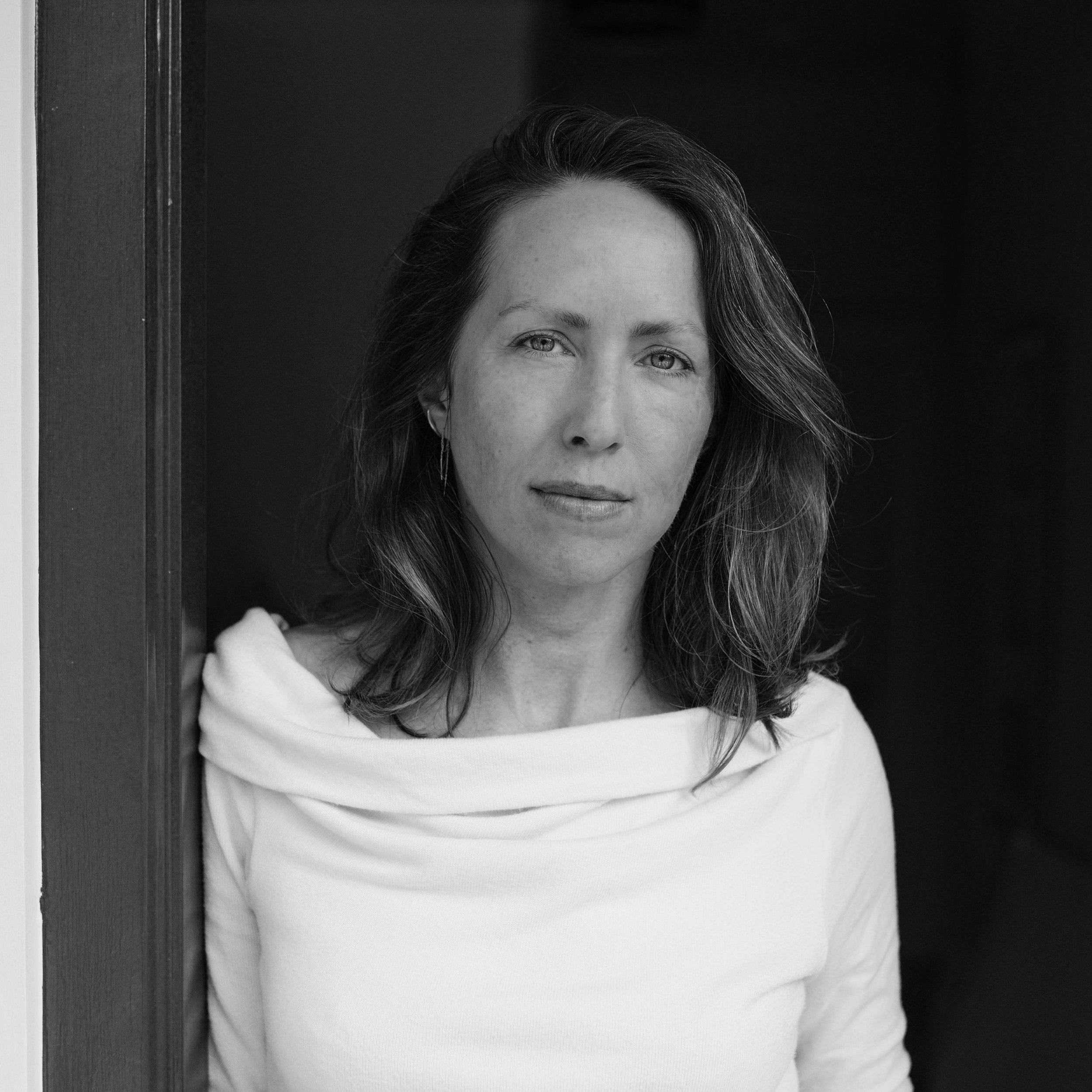 Black and white portrait of a woman with shoulder-length wavy hair, wearing a white top with a wide collar, standing near a doorway.