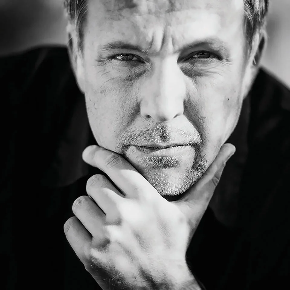 Close-up black and white photo of a man with a serious expression, resting his chin on his hand, with visible facial hair and short hair.