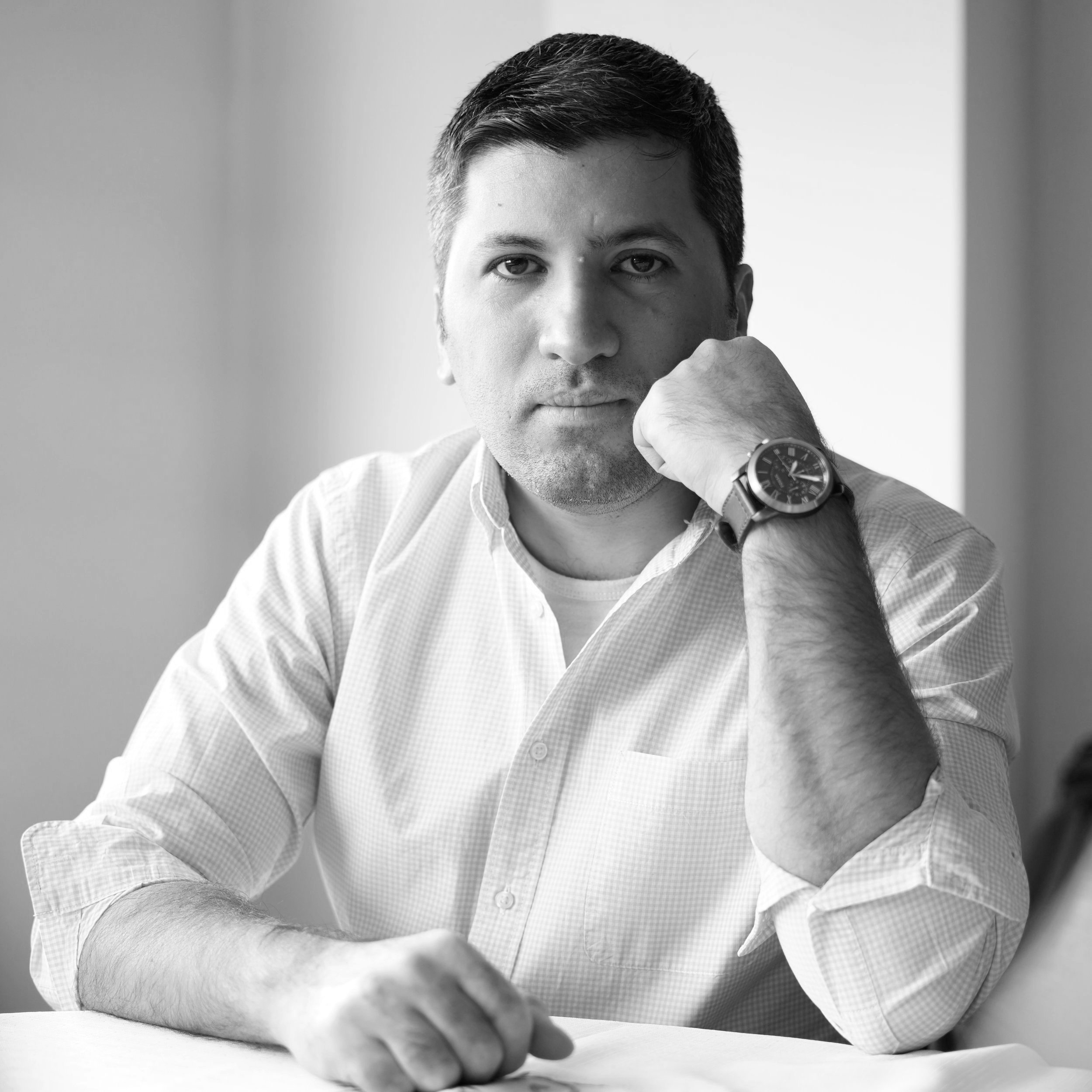 Black and white portrait of a man wearing a striped shirt, sitting at a table, resting his chin on his hand, with a watch on his wrist, looking directly at the camera.