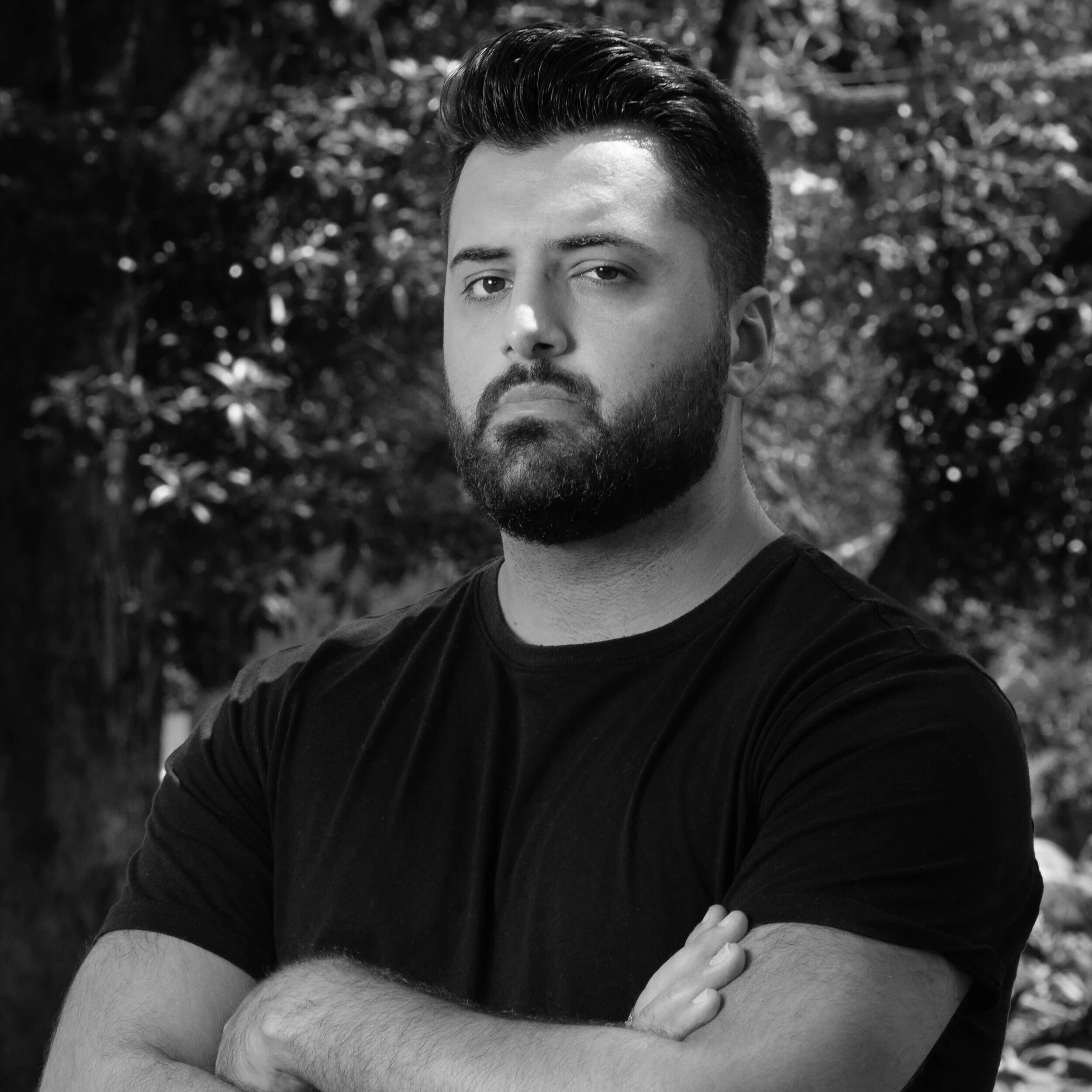 Black and white portrait of a man with short hair and a beard, wearing a black t-shirt, with arms crossed, standing outdoors in front of trees.