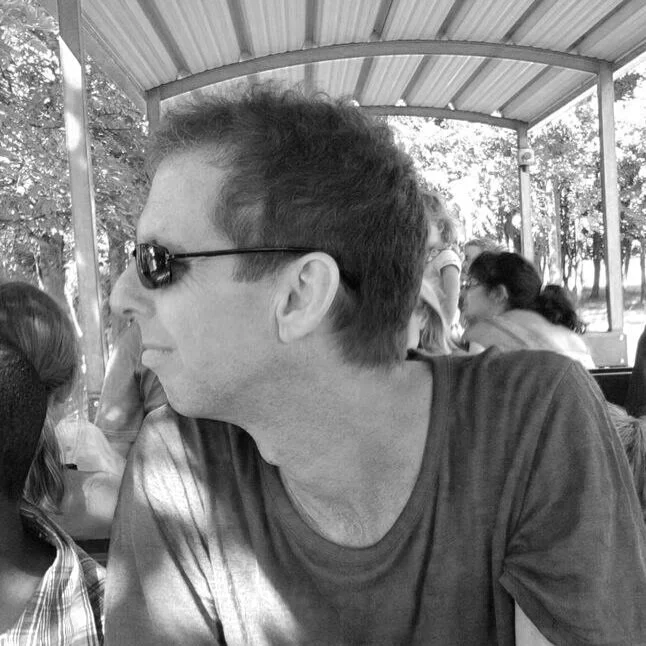 A man wearing sunglasses, sitting at a table in a covered outdoor area with other people, with trees visible outside.