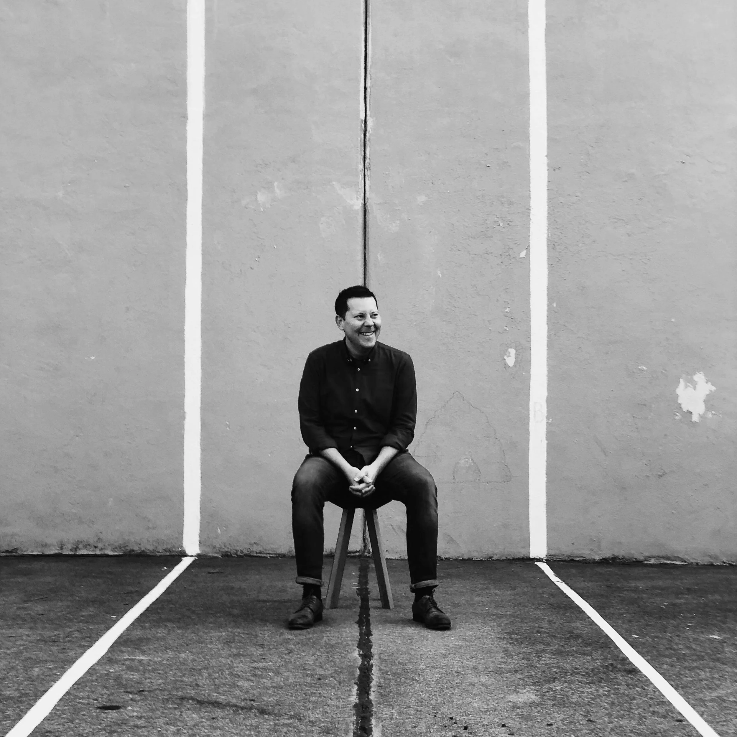 A person sitting on a stool, smiling, in front of a plain wall with sports court lines on the ground.