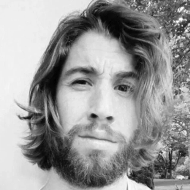 A black and white portrait of a person with long, wavy hair and a beard, looking directly at the camera with a neutral expression.