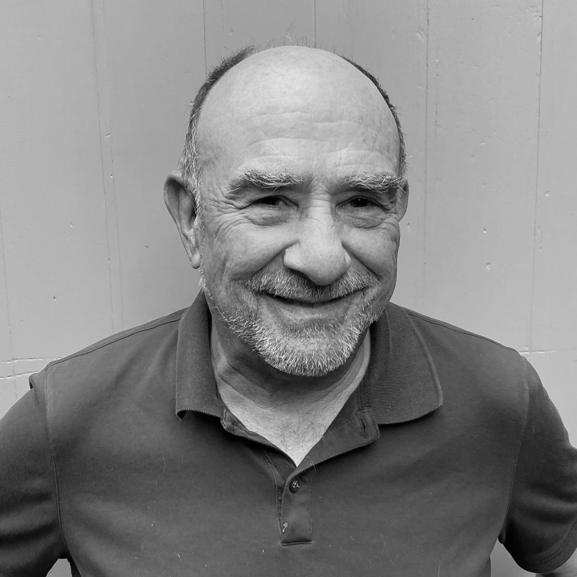 Black and white photo of smiling older man with a beard and bald head, wearing a collared shirt, against a plain wall background.