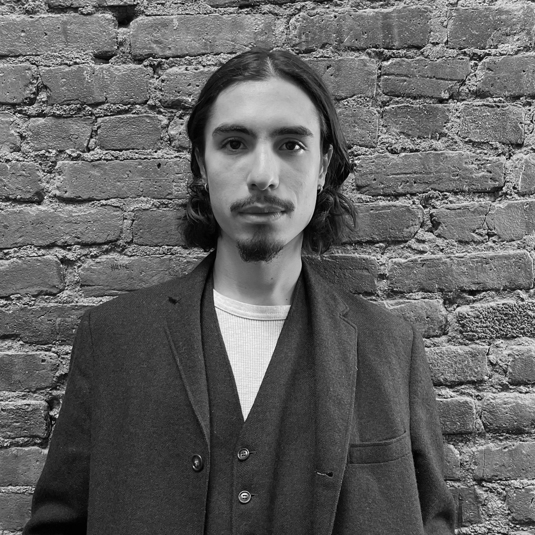 Young man with long hair, goatee, and earrings standing against a brick wall.