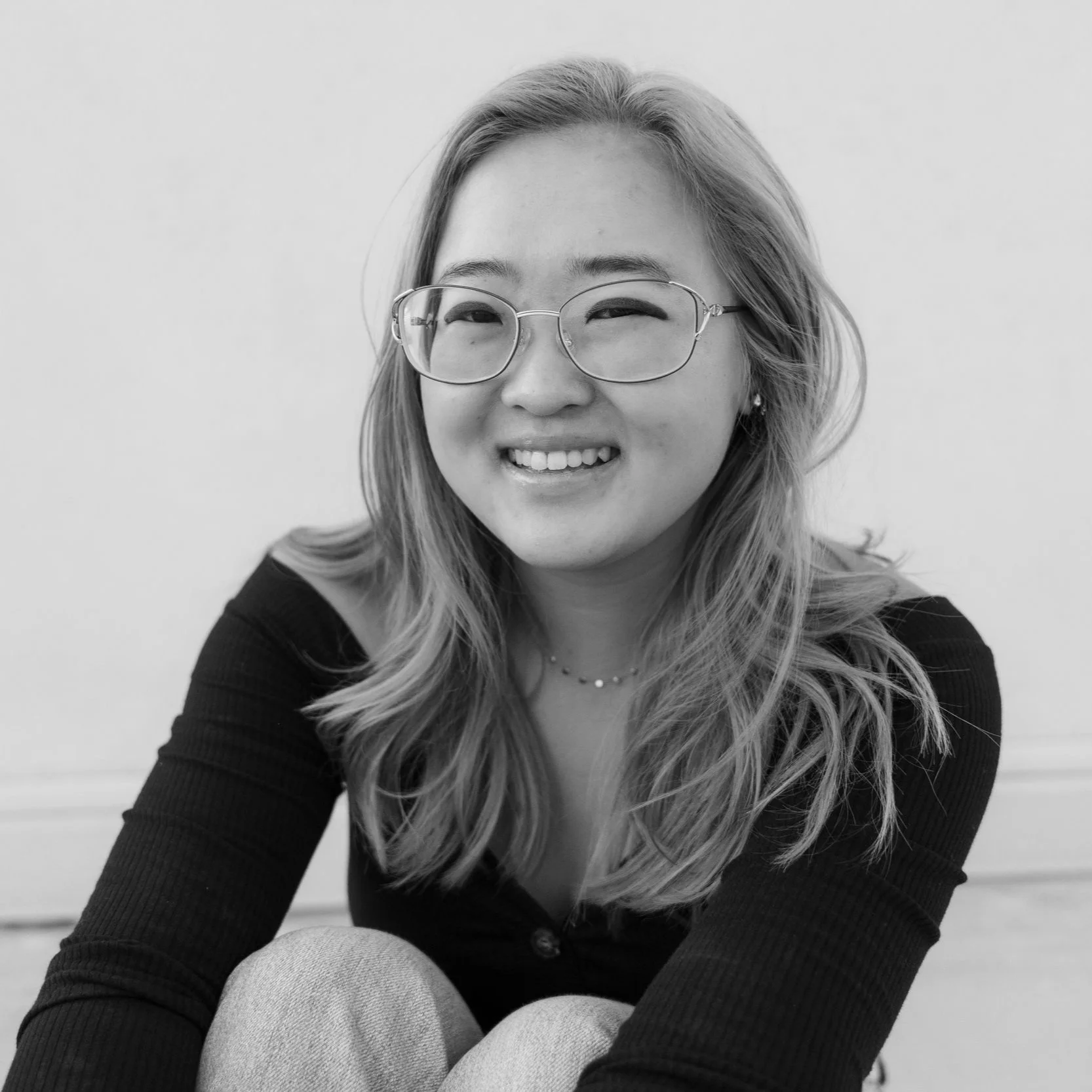 Black and white portrait of a smiling young woman with glasses, wavy hair, wearing a dark top and light-colored pants, sitting against a plain background.