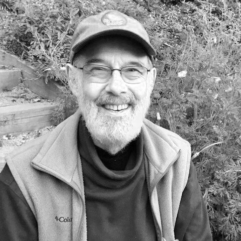 A smiling man with glasses, a beard, and a baseball cap, wearing outdoor clothing, sitting near plants or shrubs.