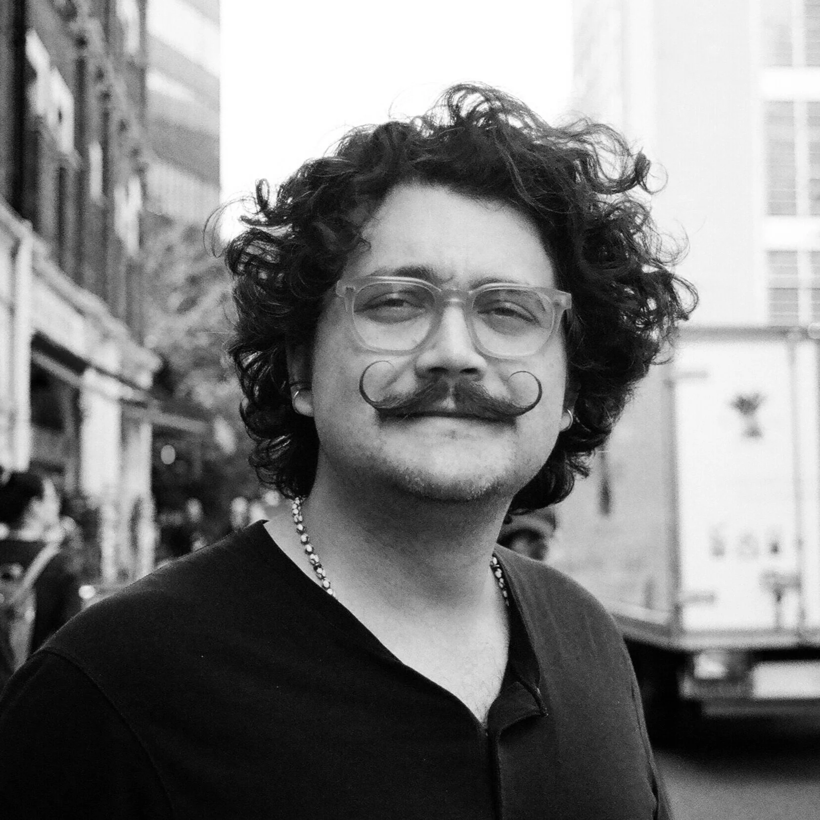 A black and white photo of a man with curly hair, glasses, a mustache with curled ends, wearing a necklace, and a dark top, standing outdoors in an urban setting.