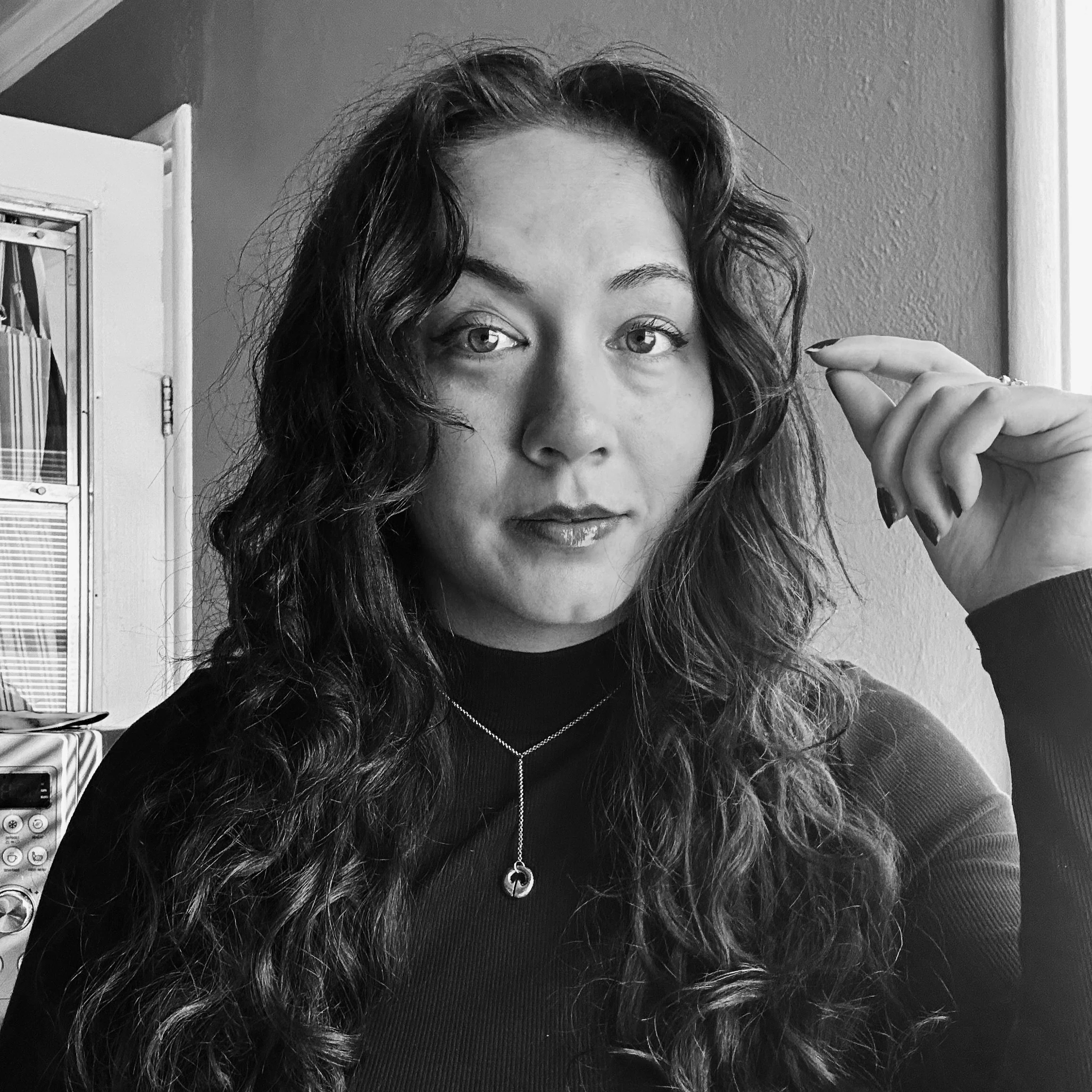 Black and white photo of a woman with long, wavy hair, wearing a black top and a necklace, looking directly at the camera, with her right hand raised near her face.