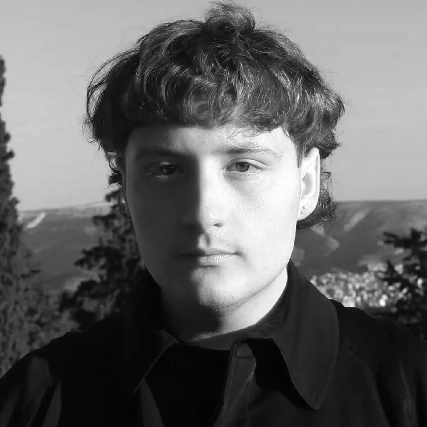 Black and white portrait of a young man with curly hair, wearing a dark shirt, standing outdoors with trees and hills in the background.