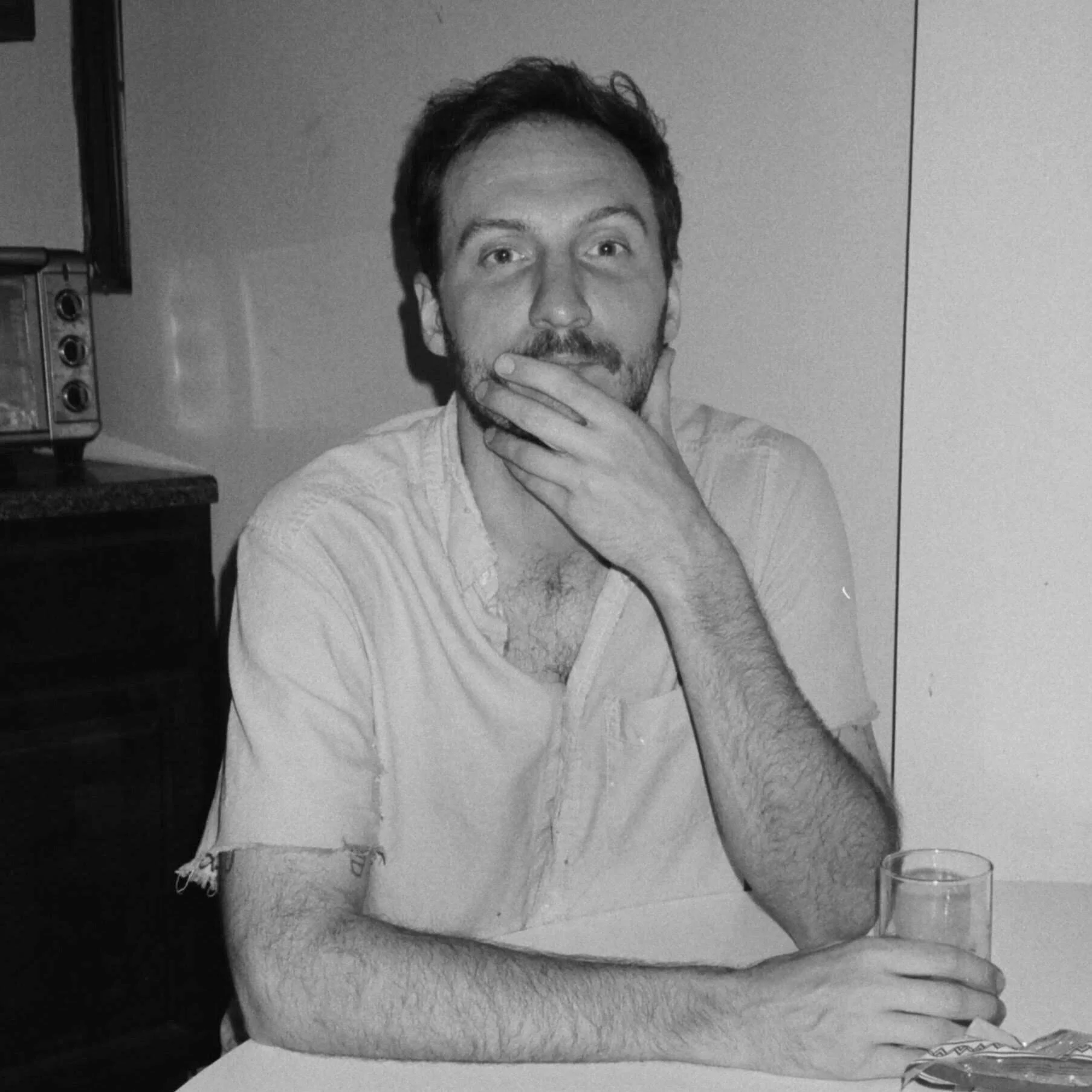 A young man with dark hair and a beard sitting at a table, holding a glass in his left hand, with his right hand resting on his chin, and looking thoughtful.