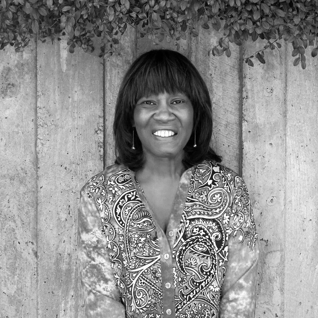 A woman smiling, wearing a patterned shirt with floral and swirl designs, standing in front of a concrete wall with foliage overhead.