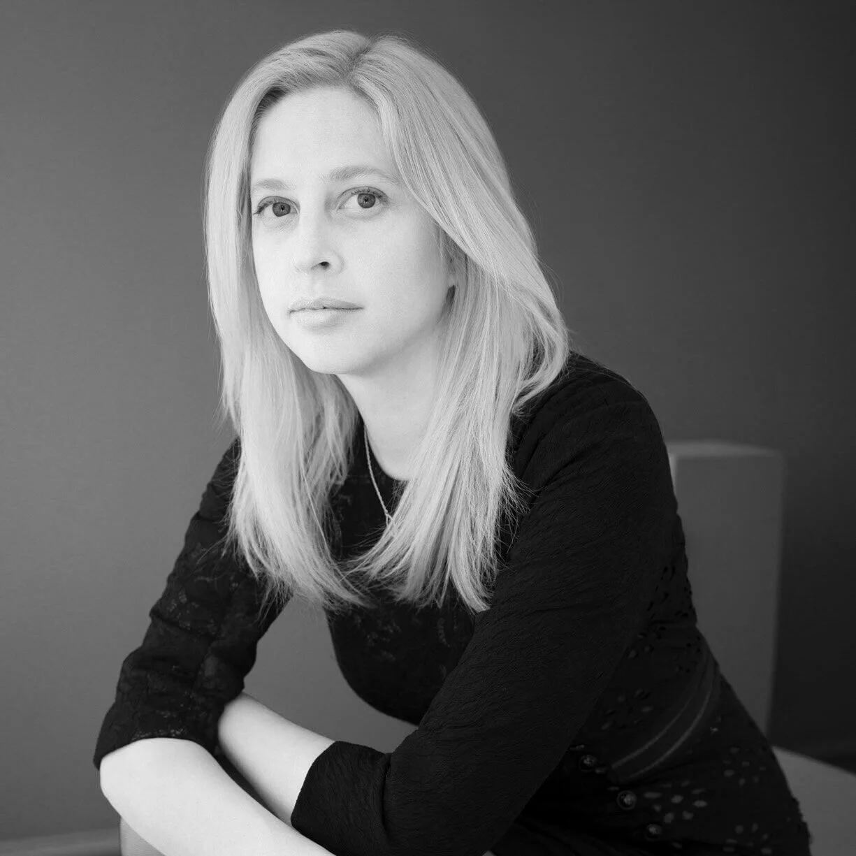Black and white portrait of a woman with straight, shoulder-length hair, wearing a black long-sleeve top, looking directly at the camera with a neutral expression, seated against a plain background.
