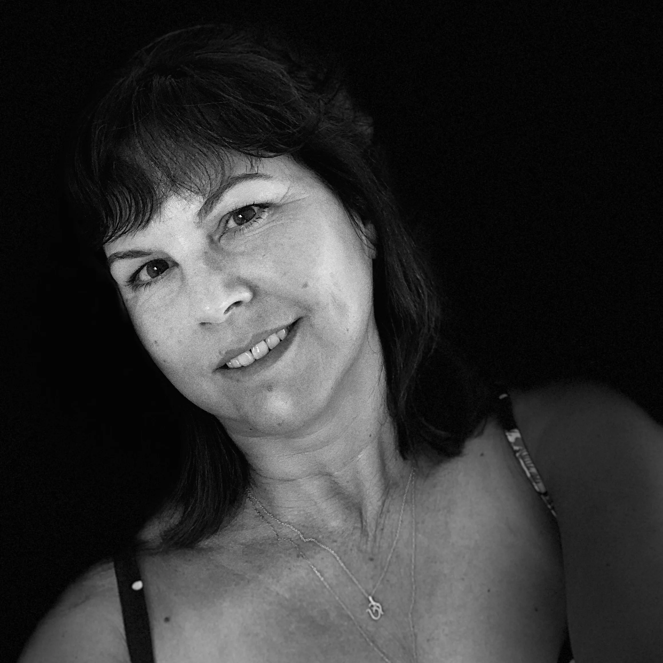 Black and white photo of a woman with dark hair, smiling, wearing a necklace and a sleeveless top, against a dark background.