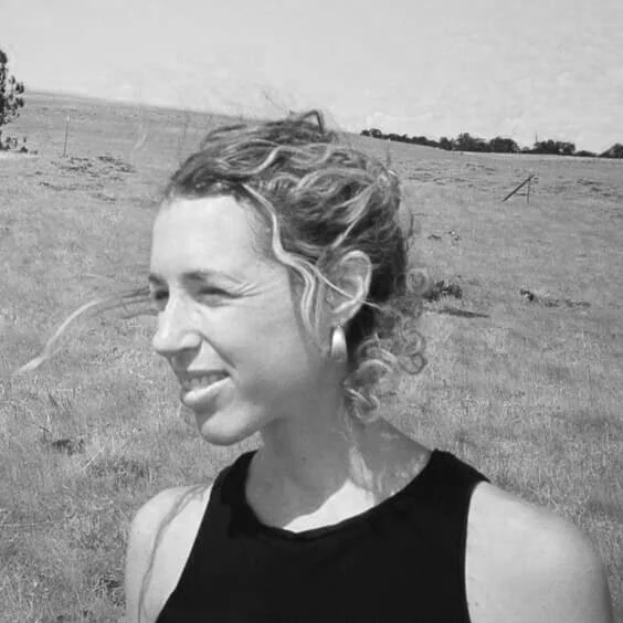 A woman with curly hair, wearing a sleeveless top, smiling outdoors in a grassy field on a windy day.