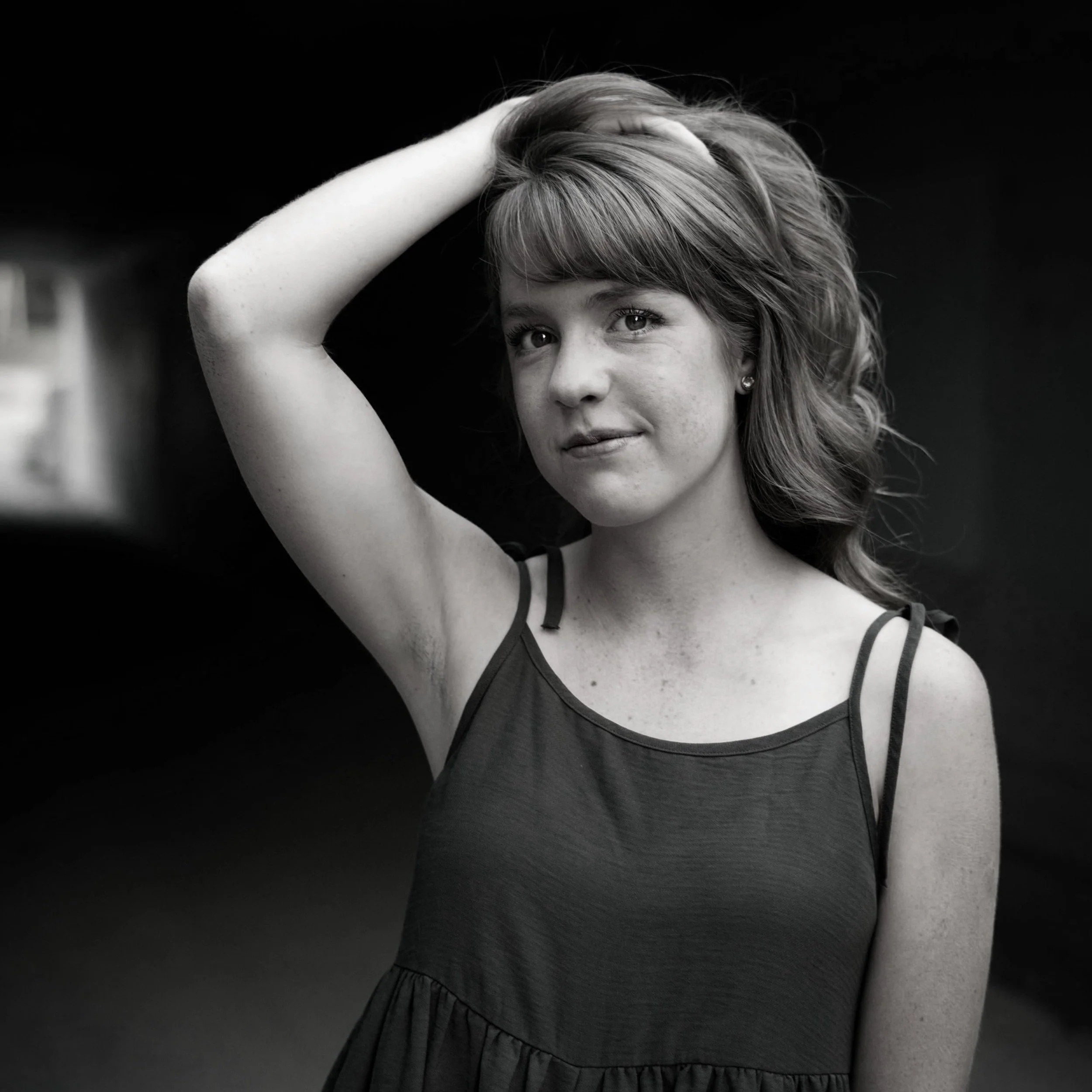 A young woman with light skin and shoulder-length wavy hair, wearing a sleeveless dress, posing with one hand on her head against a dark background.
