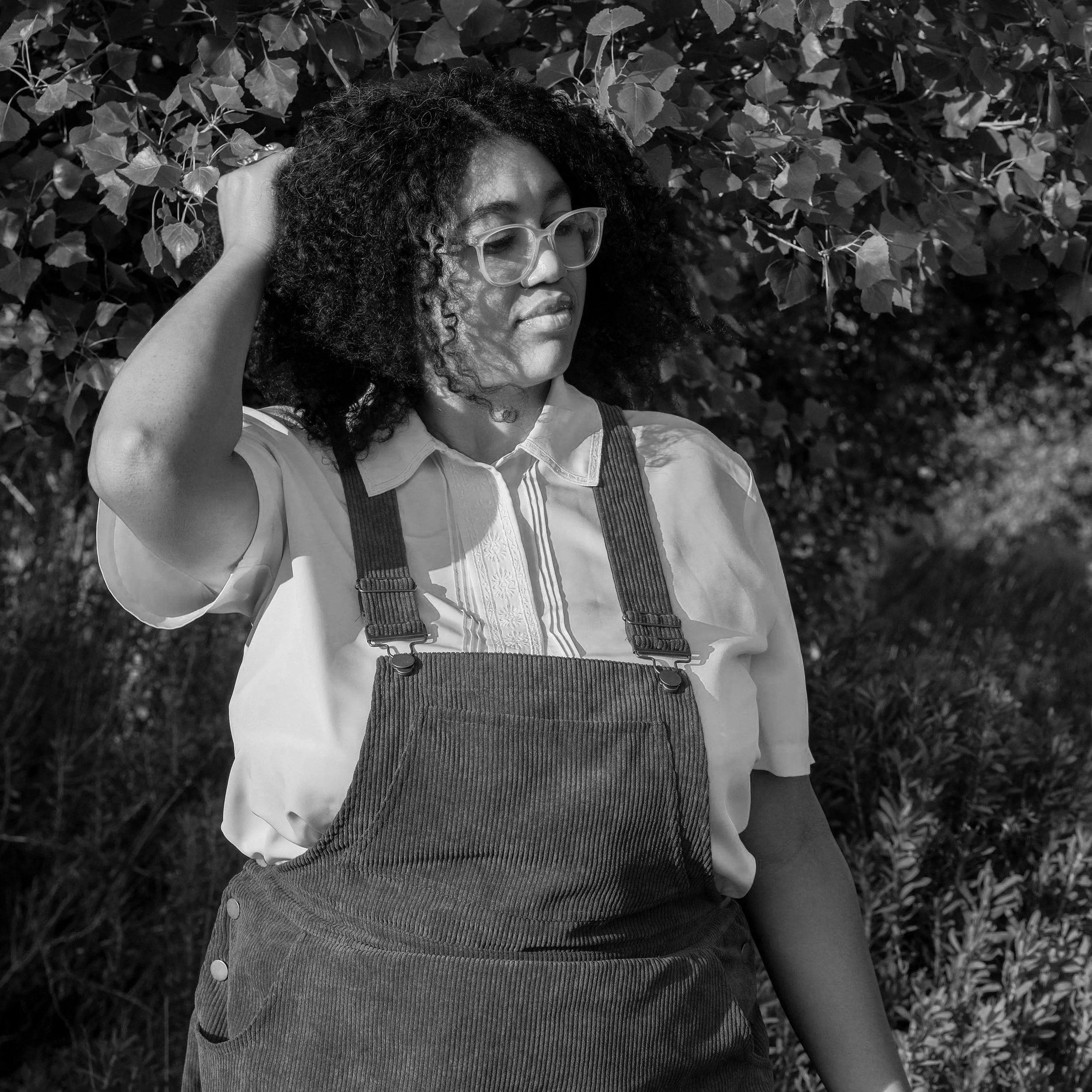 A woman with curly hair and glasses standing outdoors, touching her head with her right hand, dressed in a collared shirt and overalls, with foliage in the background.