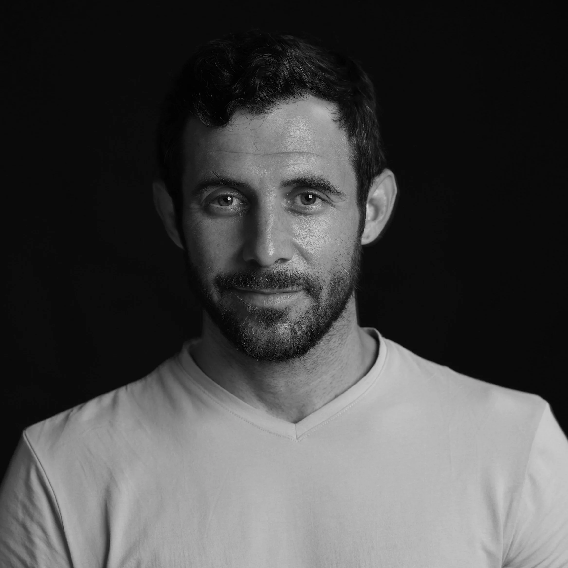 Black and white portrait of a young man with short dark hair, beard, and mustache, wearing a light-colored V-neck shirt, against a dark background.