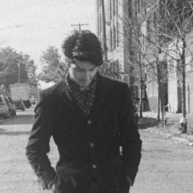A young man with dark, curly hair looking down, wearing a dark coat and patterned shirt, walking on a sidewalk in a neighborhood with trees and houses.
