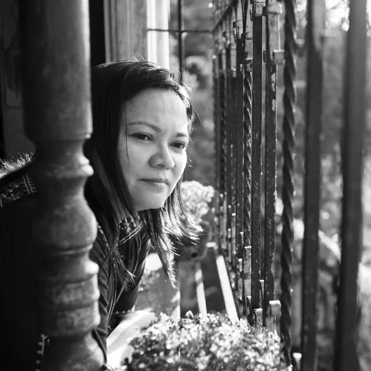 A young woman with dark hair gazing out from behind a wooden porch railing, with a black and white filter.