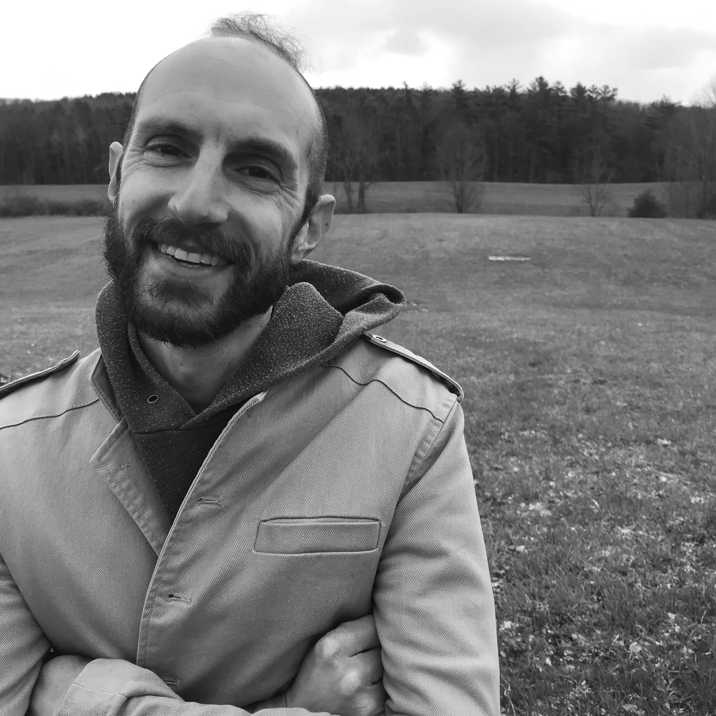 A smiling man with a beard, wearing a coat and hoodie, standing outdoors in a field with trees in the background.