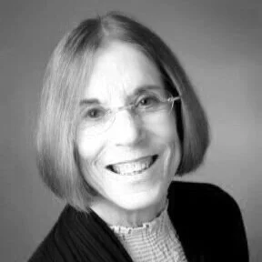 A black-and-white portrait of a smiling woman with shoulder-length hair and glasses, wearing a dark jacket over a light blouse.