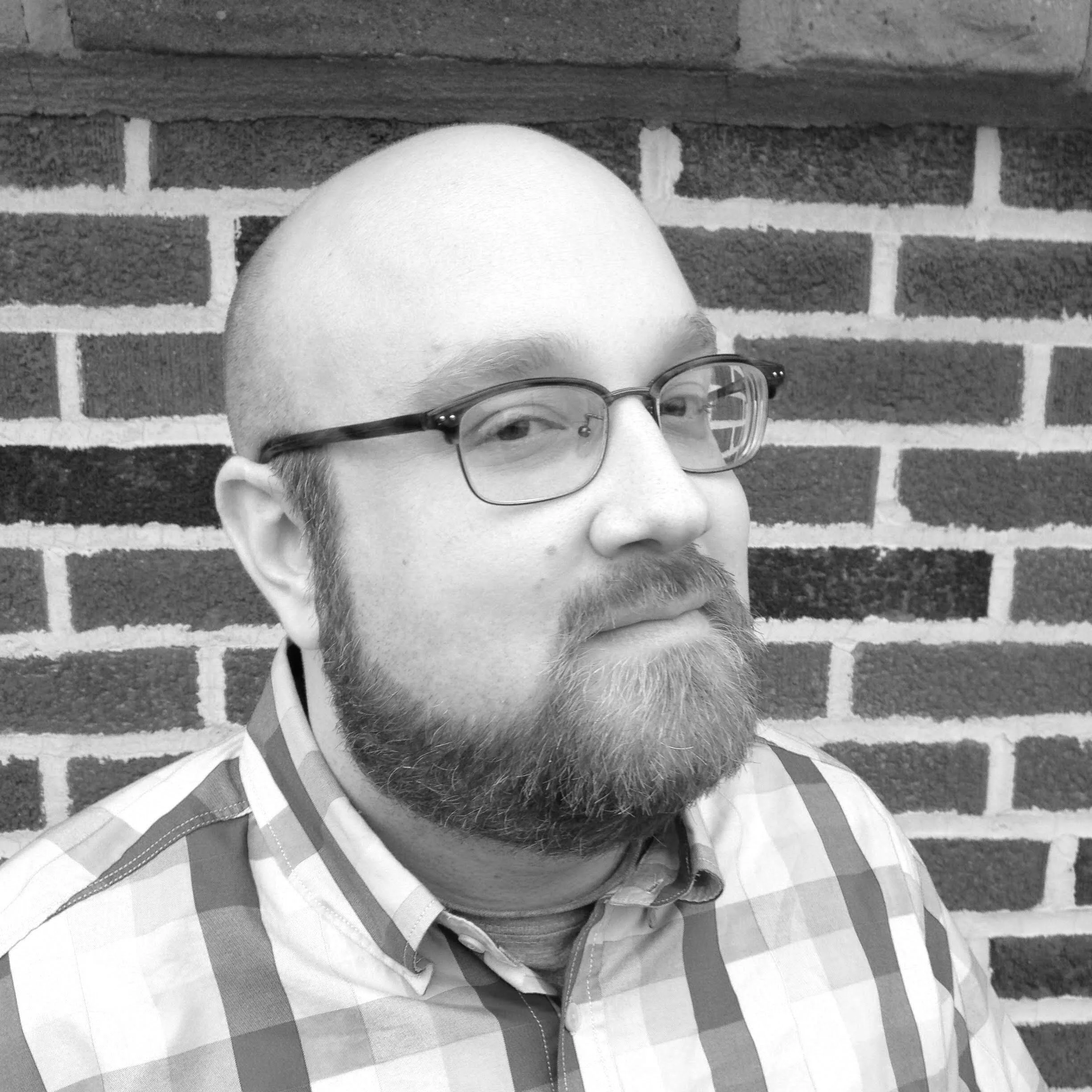 A black-and-white photo of a man with a beard and glasses, wearing a checkered shirt, standing against a brick wall.