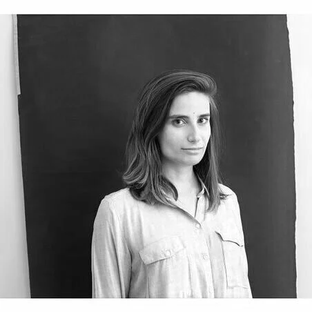 Black and white photo of a young woman with shoulder-length hair, wearing a light-colored button-up shirt, standing in front of a dark background.