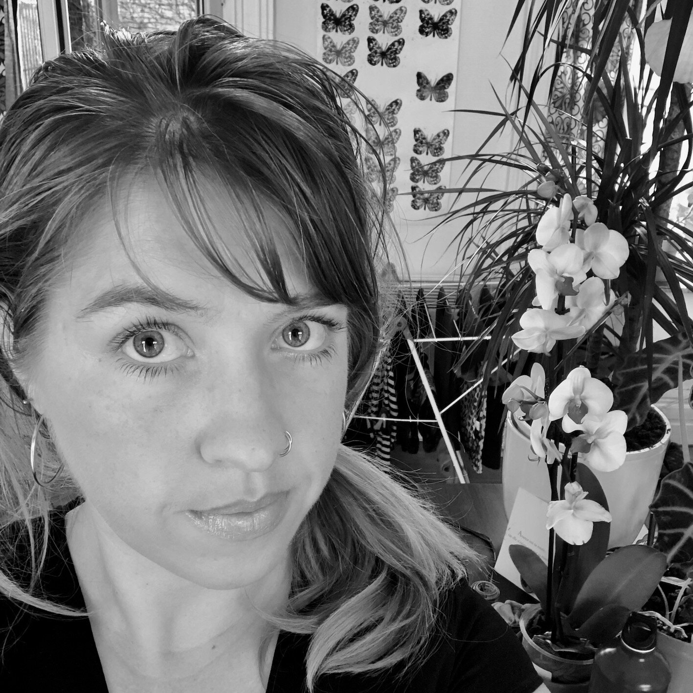 A black and white photo of a young woman with light-colored eyes, wearing earrings and a nose ring, standing near a flowering orchid with large leaves, in an indoor setting decorated with butterfly artwork on the wall.