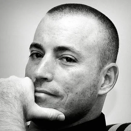 Close-up of a man with short hair resting his chin on his hand, looking at the camera in black and white.