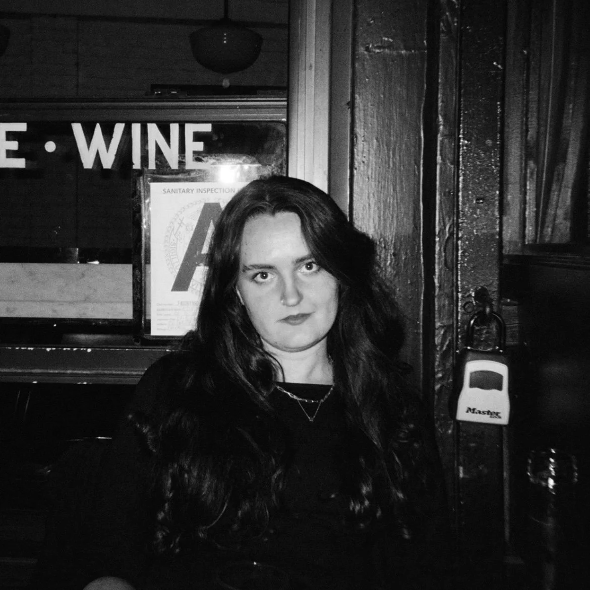A woman with long dark hair and a nose ring sitting indoors near a wooden wall, with a sign that reads 'WINE' and a sanitation inspection sheet behind her.