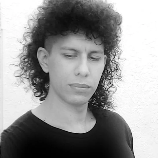 Black and white photo of a person with curly hair and a serious expression, looking down, wearing a black shirt, standing against a plain light-colored wall.