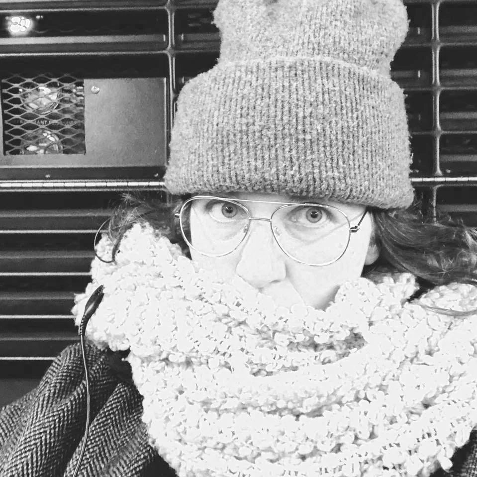 A person wearing glasses, a knit beanie, and a scarf, sitting in front of a vent.