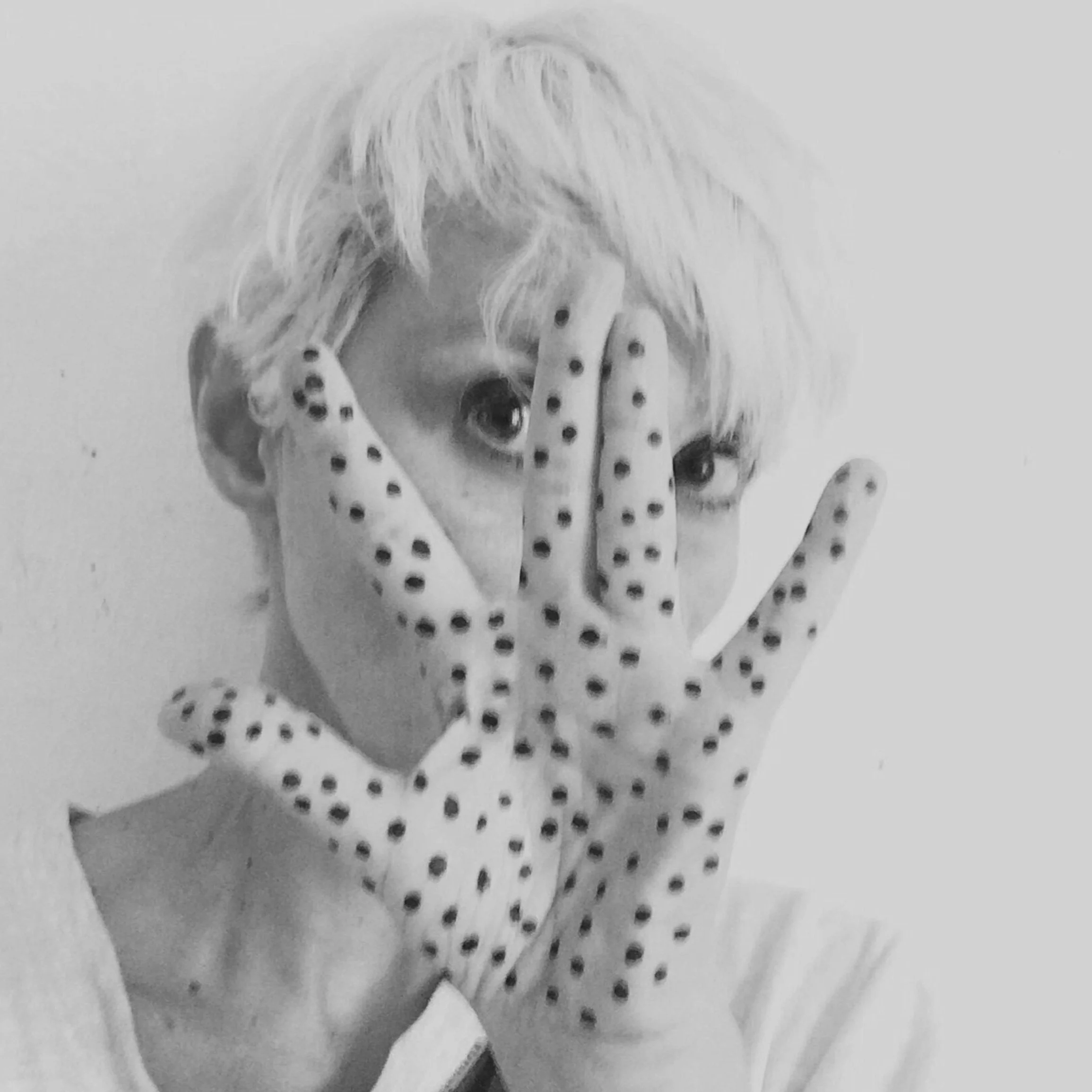 A person with short, tousled light hair holding a polka-dotted glove in front of their face, covering their nose and mouth, with visible eyes peeking through the glove.