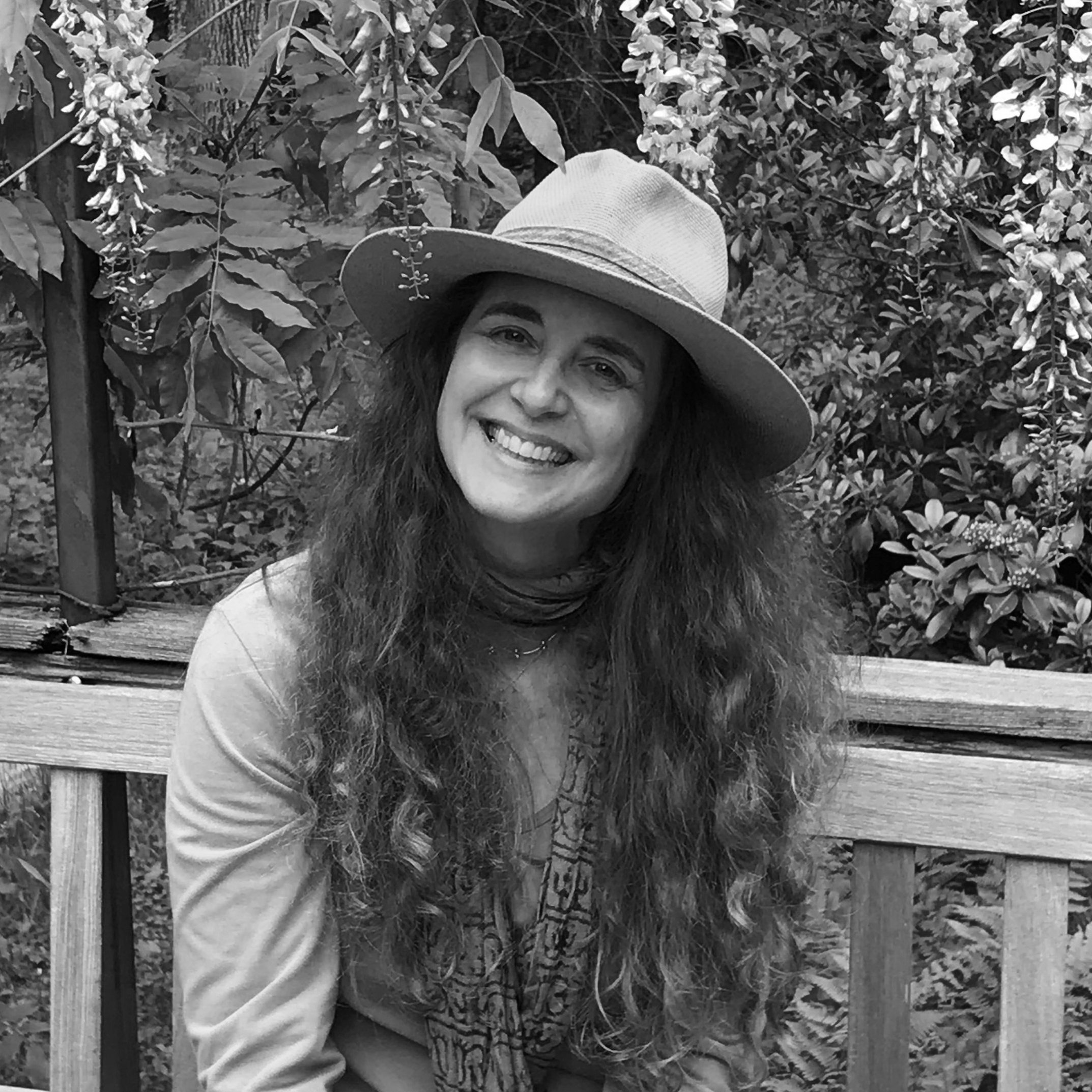 A smiling woman with long curly hair wearing a wide-brimmed hat and a scarf, sitting on a wooden bench outdoors surrounded by plants and flowers.