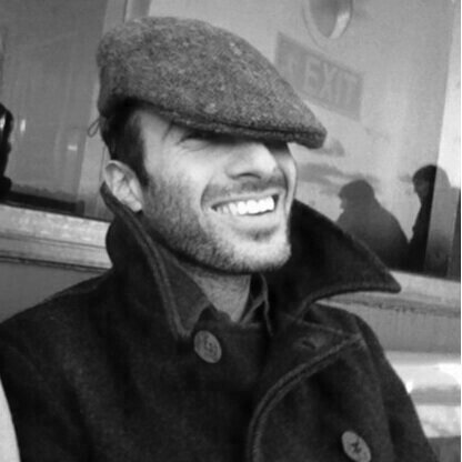 Smiling man wearing a flat cap and a dark coat, sitting indoors.