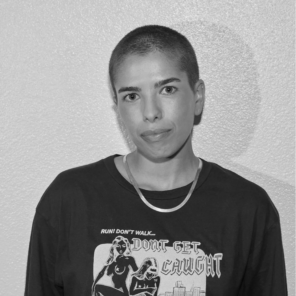 A person with short hair wearing a black T-shirt with a graphic design and text, standing against a textured wall.