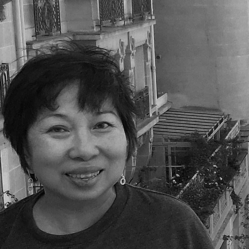 Black and white photo of a smiling woman with short dark hair, wearing earrings, in front of a Parisian-style street with balconies and plants.