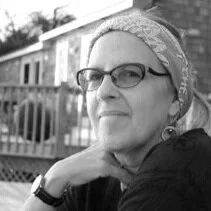 Black-and-white photo of a woman with glasses and a headscarf, outdoors near a house.
