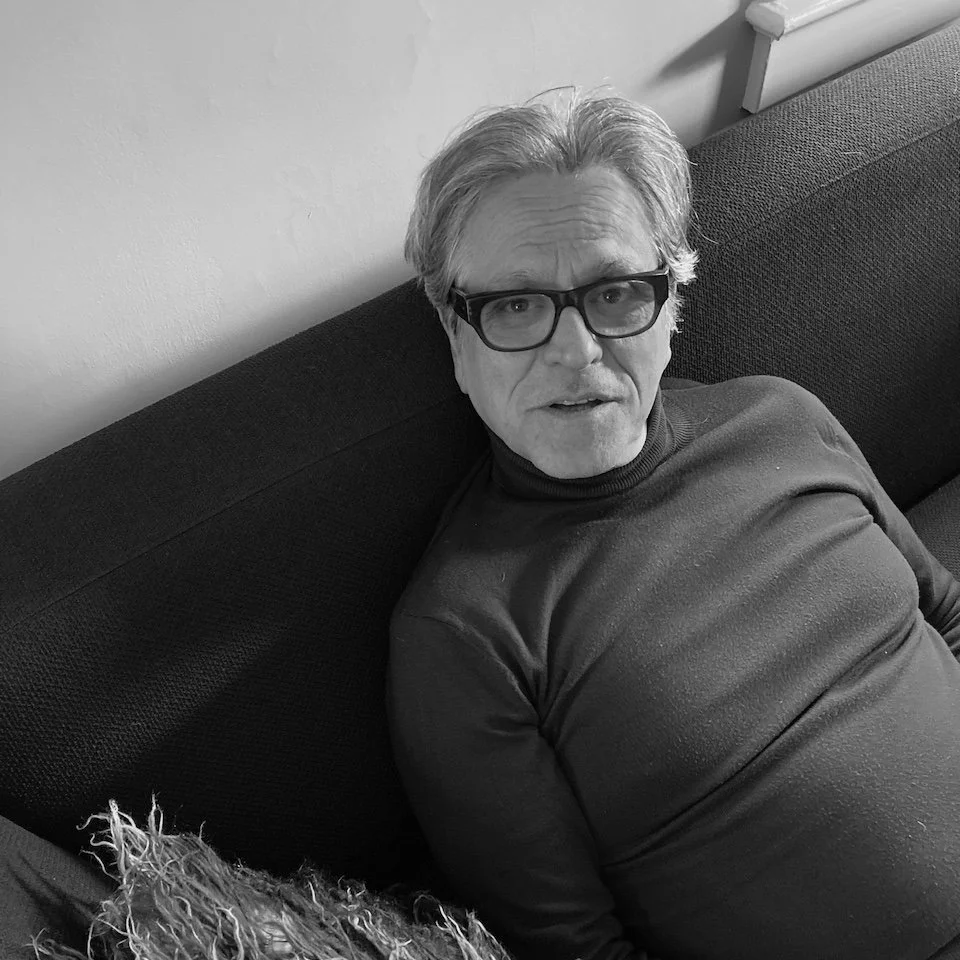 A man with gray hair and glasses sitting on a dark-colored sofa, looking up at the camera