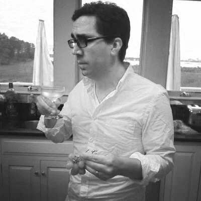 Black and white photo of a man with glasses and dark hair holding a glass and a plate with food in a kitchen with windows and curtains in the background.