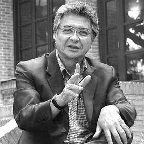 A woman with short, curly hair and glasses, wearing a blazer, making a peace sign with her right hand in front of a brick building.