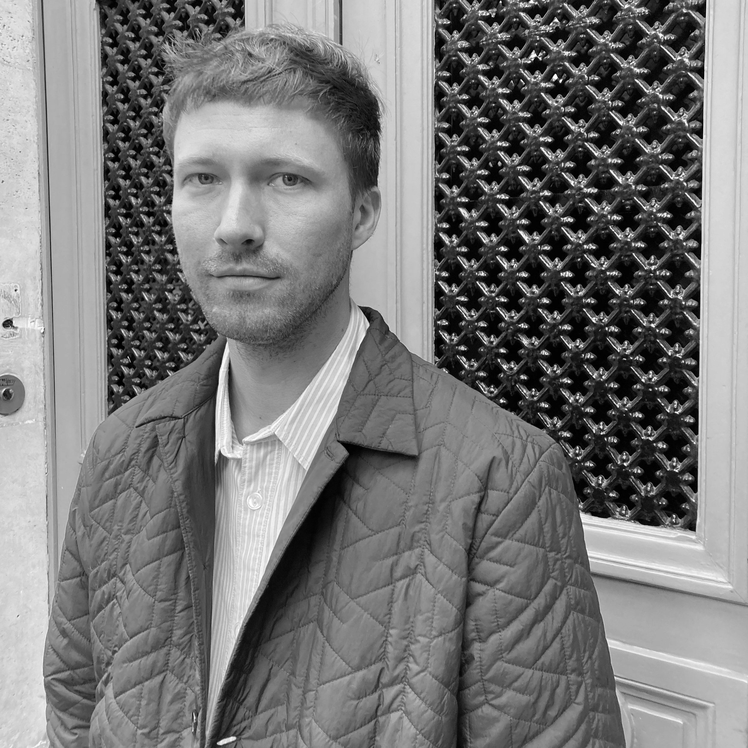 A man with short hair and light skin in a quilted jacket and collared shirt standing in front of a decorative metal grate.